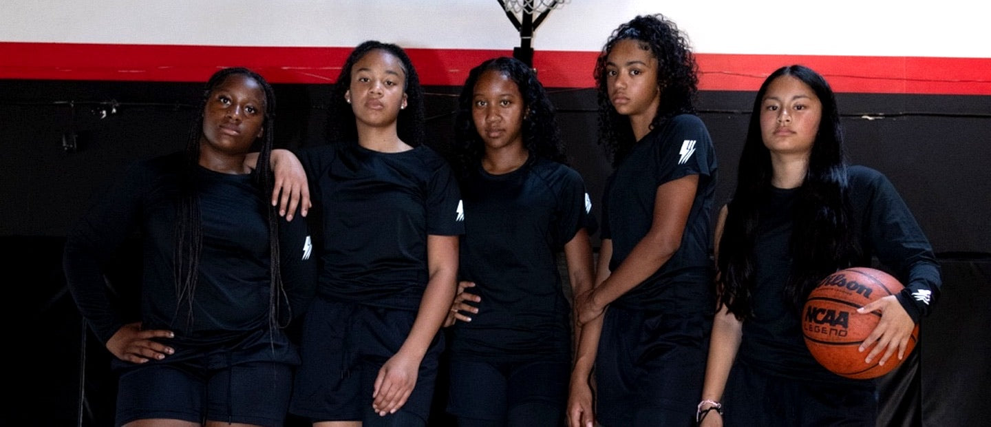 Five young women in black athletic wear standing together with a basketball against a red and black background.