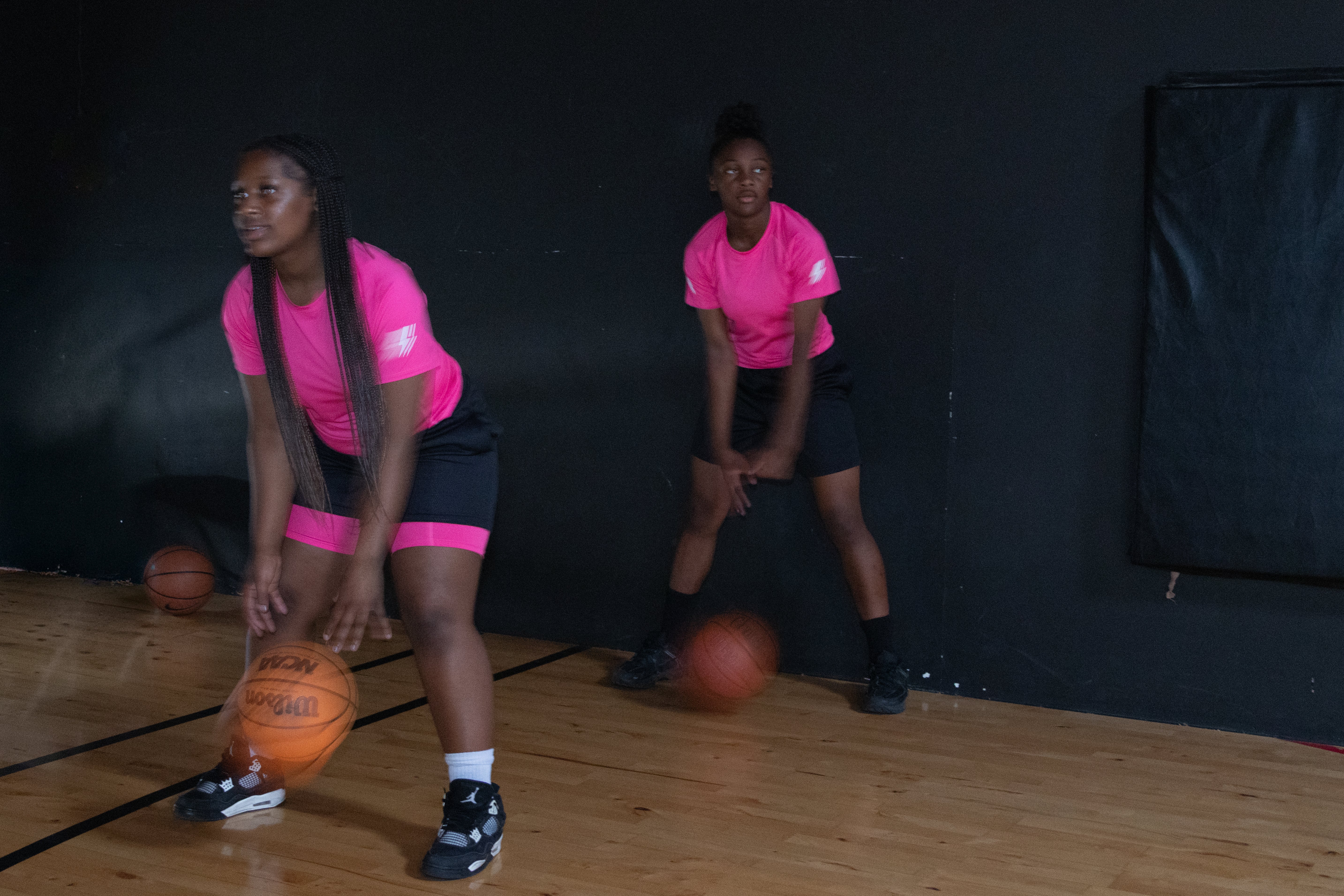 Two women in pink shirts and black shorts holding basketballs on a wooden floor with a dark background.